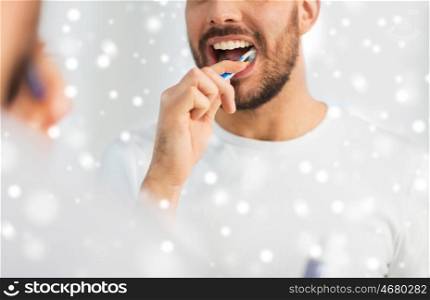 health care, dental hygiene, people and beauty concept - close up of young man with toothbrush cleaning teeth and looking to mirror at home bathroom over snow
