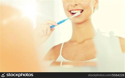 health care, dental hygiene, people and beauty concept - close up of smiling young woman with toothbrush cleaning teeth and looking to mirror at home bathroom. woman with toothbrush cleaning teeth at bathroom. woman with toothbrush cleaning teeth at bathroom