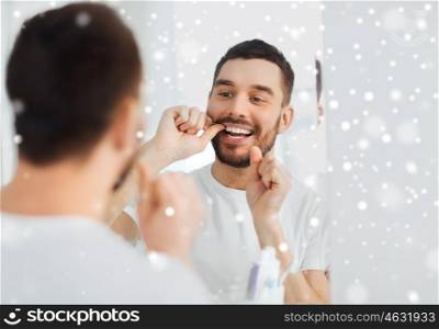health care, dental hygiene, people and beauty concept - smiling young man with floss cleaning teeth and looking to mirror at home bathroom over snow