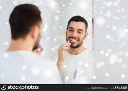 health care, dental hygiene, people and beauty concept - smiling young man with toothbrush cleaning teeth and looking to mirror at home bathroom over snow