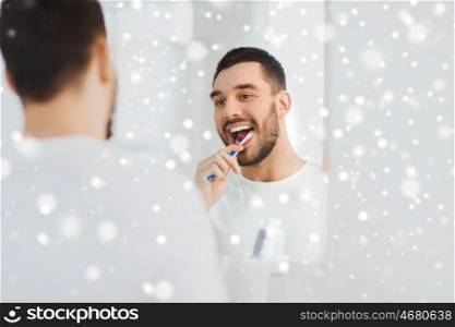 health care, dental hygiene, people and beauty concept - smiling young man with toothbrush cleaning teeth and looking to mirror at home bathroom over snow
