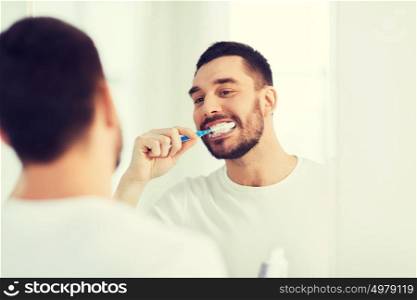health care, dental hygiene, people and beauty concept - smiling young man with toothbrush cleaning teeth and looking to mirror at home bathroom. man with toothbrush cleaning teeth at bathroom