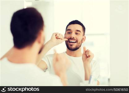 health care, dental hygiene, people and beauty concept - smiling young man with floss cleaning teeth and looking to mirror at home bathroom. man with dental floss cleaning teeth at bathroom