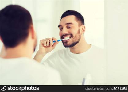 health care, dental hygiene, people and beauty concept - smiling young man with toothbrush cleaning teeth and looking to mirror at home bathroom. man with toothbrush cleaning teeth at bathroom