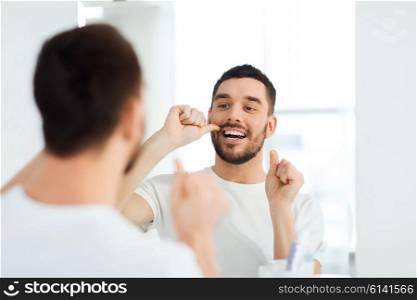 health care, dental hygiene, people and beauty concept - smiling young man with floss cleaning teeth and looking to mirror at home bathroom