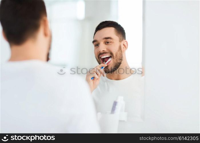 health care, dental hygiene, people and beauty concept - smiling young man with toothbrush cleaning teeth and looking to mirror at home bathroom