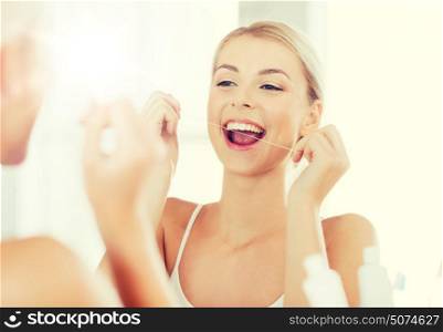 health care, dental hygiene, people and beauty concept - smiling young woman with floss cleaning teeth and looking to mirror at home bathroom. woman with dental floss cleaning teeth at bathroom
