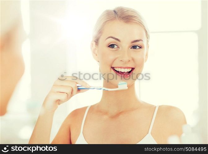 health care, dental hygiene, people and beauty concept - smiling young woman with toothbrush cleaning teeth and looking to mirror at home bathroom. woman with toothbrush cleaning teeth at bathroom. woman with toothbrush cleaning teeth at bathroom