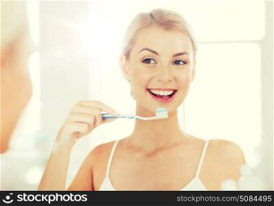 health care, dental hygiene, people and beauty concept - smiling young woman with toothbrush cleaning teeth and looking to mirror at home bathroom. woman with toothbrush cleaning teeth at bathroom. woman with toothbrush cleaning teeth at bathroom