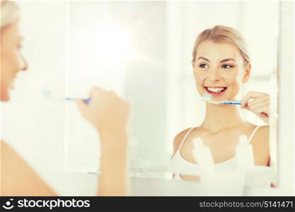 health care, dental hygiene, people and beauty concept - smiling young woman with toothbrush cleaning teeth and looking to mirror at home bathroom. woman with toothbrush cleaning teeth at bathroom