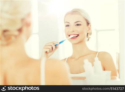 health care, dental hygiene, people and beauty concept - smiling young woman with toothbrush cleaning teeth and looking to mirror at home bathroom. woman with toothbrush cleaning teeth at bathroom