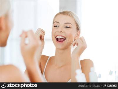 health care, dental hygiene, people and beauty concept - smiling young woman with floss cleaning teeth and looking to mirror at home bathroom