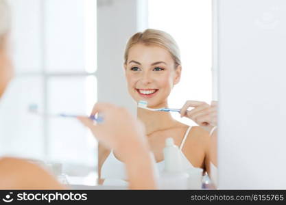 health care, dental hygiene, people and beauty concept - smiling young woman with toothbrush cleaning teeth and looking to mirror at home bathroom