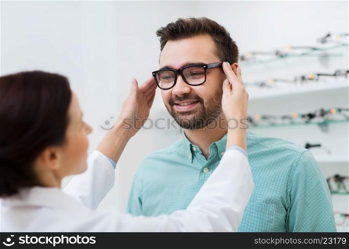 health care, people, eyesight and vision concept - female optician putting on glasses to man at optics store
