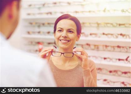 health care, people, eyesight and vision concept - happy woman choosing and showing glasses to optician at optics store