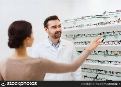 health care, people, eyesight and vision concept - happy woman choosing and showing glasses to optician at optics store