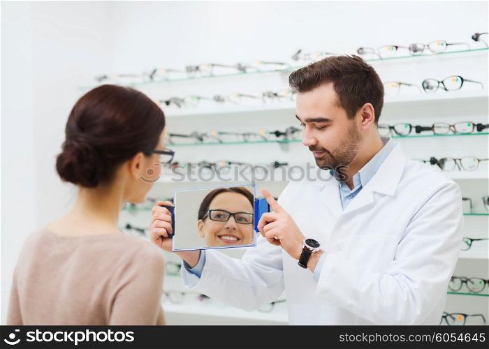 health care, people, eyesight and vision concept - optician and woman in glasses looking to mirror at optics store