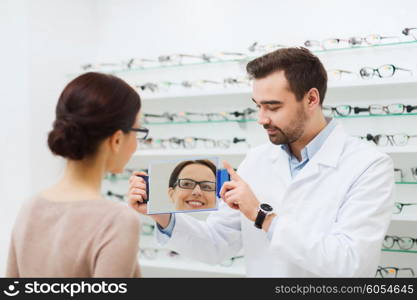 health care, people, eyesight and vision concept - optician and woman in glasses looking to mirror at optics store
