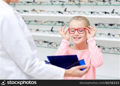 health care, people, eyesight and vision concept - optician with mirror helping little girl to choose glasses at optics store