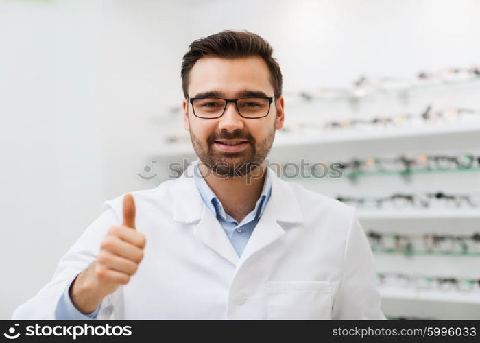 health care, people, eyesight and vision concept - smiling man optician in glasses and white coat showing thumbs up gesture at optics store