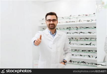 health care, people, eyesight and vision concept - smiling man optician in glasses and white coat showing thumbs up gesture at optics store