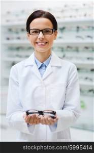 health care, people, eyesight and vision concept - smiling woman optician holding glasses at optics store
