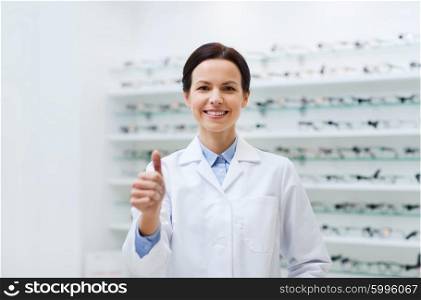 health care, people, eyesight and vision concept - smiling woman optician in white coat showing thumbs up gesture over glasses at optics store