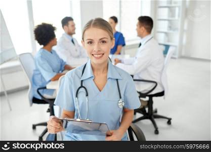 health care, profession, people and medicine concept - happy female doctor or nurse with clipboard over group of medics meeting at hospital