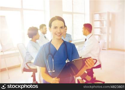 health care, profession, people and medicine concept - happy female doctor with clipboard over group of medics meeting at hospital
