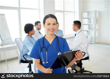 health care, profession, people and medicine concept - happy female doctor with clipboard over group of medics meeting at hospital