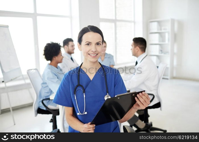 health care, profession, people and medicine concept - happy female doctor with clipboard over group of medics meeting at hospital