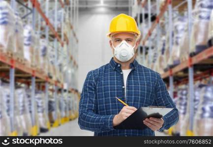 health care, work and pandemic concept - male worker in safety helmet wearing face protective mask or respirator with clipboard and pencil over warehouse background. male worker in mask with clipboard at warehouse
