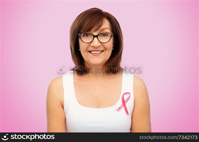 health, charity and old people concept - portrait of smiling senior woman in glasses with breast cancer awareness ribbon over pink background. old woman with pink breast cancer awareness ribbon