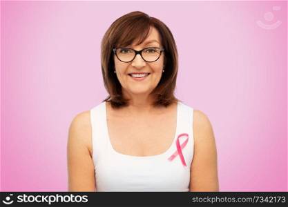 health, charity and old people concept - portrait of smiling senior woman in glasses with breast cancer awareness ribbon over pink background. old woman with pink breast cancer awareness ribbon