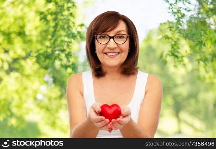 health, charity and valentine&rsquo;s day concept - portrait of smiling senior woman holding red heart over green natural background. portrait of smiling senior woman holding red heart