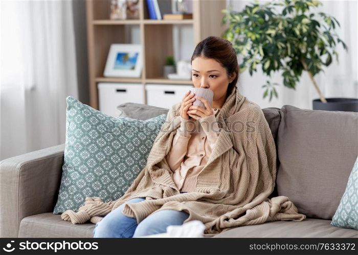 health, cold and people concept - sad sick young asian woman in blanket drinking hot tea at home. sad sick asian woman drinking hot tea at home