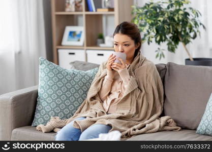 health, cold and people concept - sad sick young asian woman in blanket drinking hot tea at home. sad sick asian woman drinking hot tea at home