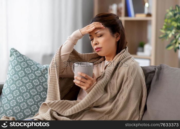 health, cold and people concept - sad sick young asian woman in blanket drinking hot tea at home. ill asian woman with headache drinking tea at home