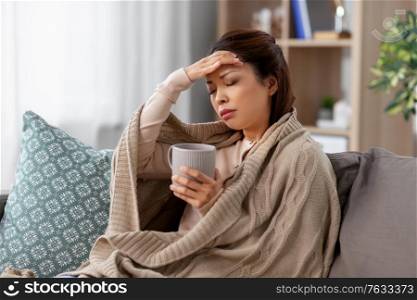 health, cold and people concept - sad sick young asian woman in blanket drinking hot tea at home. ill asian woman with headache drinking tea at home