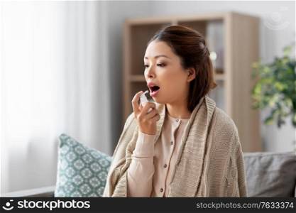 health, cold and people concept - sad sick young asian woman in blanket using oral spray medicine at home. sick asian woman using oral spray medicine at home