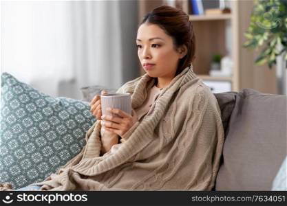 health, cold and people concept - sad sick young asian woman in blanket drinking hot tea at home. sad sick asian woman drinking hot tea at home