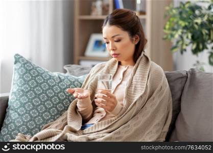 health, cold and people concept - sad sick young asian woman in blanket with medicine and glass of water at home. sick asian woman with medicine and water at home