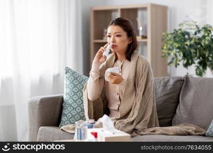 health, cold and people concept - sad sick young asian woman in blanket using nasal spray medicine and paper tissue at home. sick asian woman with nasal spray medicine at home
