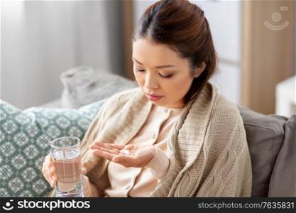 health, cold and people concept - sad sick young asian woman in blanket with medicine and glass of water at home. sick asian woman with medicine and water at home
