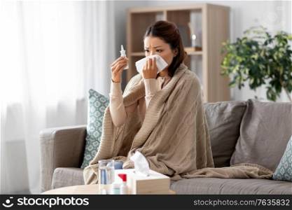 health, cold and people concept - sad sick young asian woman in blanket with nasal spray medicine and paper tissue at home. sick asian woman with nasal spray medicine at home