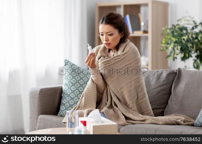 health, cold and people concept - sad sick young asian woman in blanket with nasal spray medicine at home. sick asian woman with nasal spray medicine at home