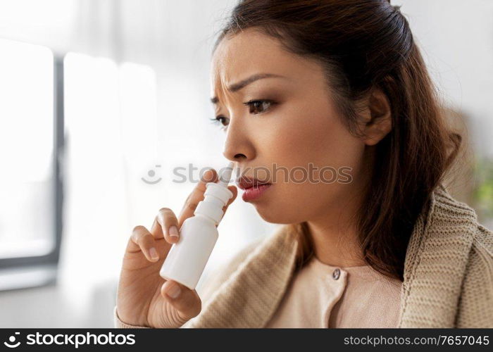 health, cold and people concept - sad sick young asian woman in blanket using nasal spray at home. sick asian woman with nasal spray medicine at home