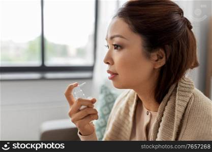 health, cold and people concept - sad sick young asian woman in blanket using oral spray medicine at home. sick asian woman using oral spray medicine at home