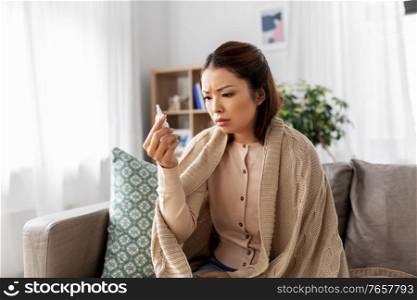 health, cold and people concept - sad sick young asian woman in blanket with nasal spray medicine at home. sick asian woman with nasal spray medicine at home