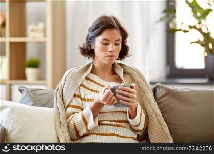 health, cold and people concept - sad sick young woman in blanket sitting on sofa and drinking hot tea at home. sad sick young woman drinking hot tea at home